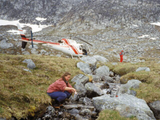 Researchers collect sediments from a rocky stream with a helicopter and steep rock hills in the background